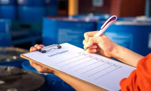 Safety inspection on the checklist document action with blurred background of chemical storage area at factory. Industrial safety concept photo. Selective focus at human's hand part.